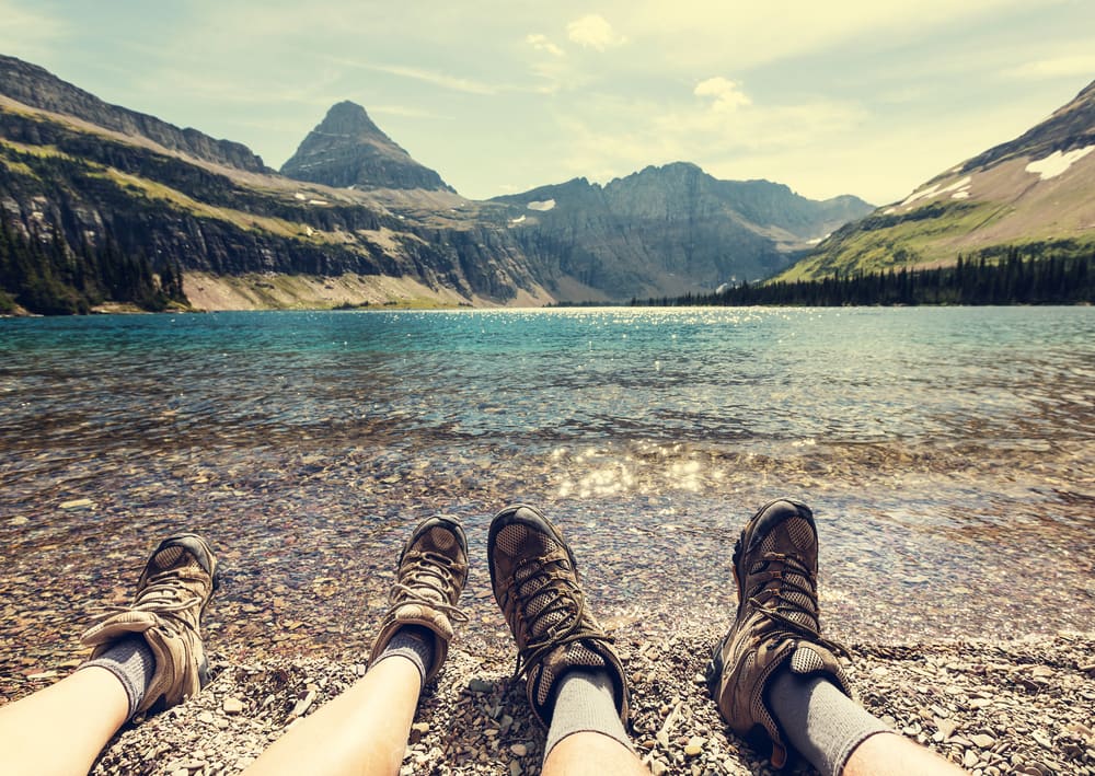 feet of two hikers sitting at the edge of the water looking at mountains